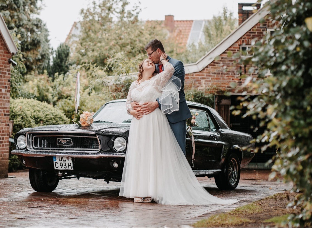 Bräutigam küsst Braut auf die Stirn neben einem Ford Mustang