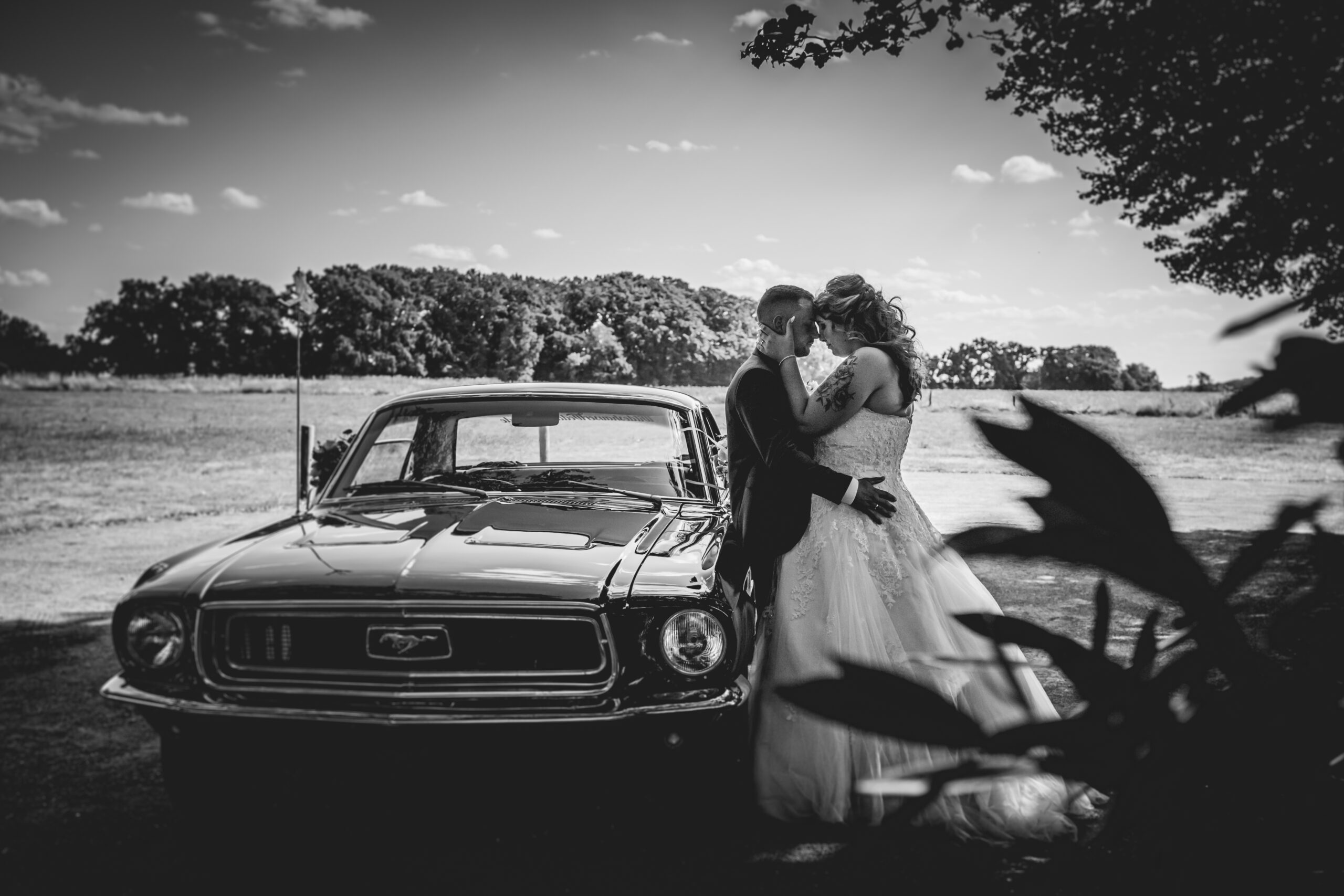 Brautpaar steht rechts neben einen Ford Mustang und guckt sich liebevoll an